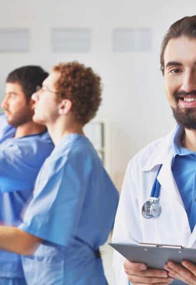 Portrait of friendly doctor looking at camera on background of his colleagues interacting